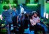 PGL Masters Bucharest 2026 Preview: Teams, Format, and Schedule Gamers interacting at a gaming lounge, with one player raising a hand for a high-five as others sit at their gaming PCs under neon green and blue lighting.