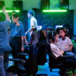 PGL Masters Bucharest 2026 Preview: Teams, Format, and Schedule Gamers interacting at a gaming lounge, with one player raising a hand for a high-five as others sit at their gaming PCs under neon green and blue lighting.